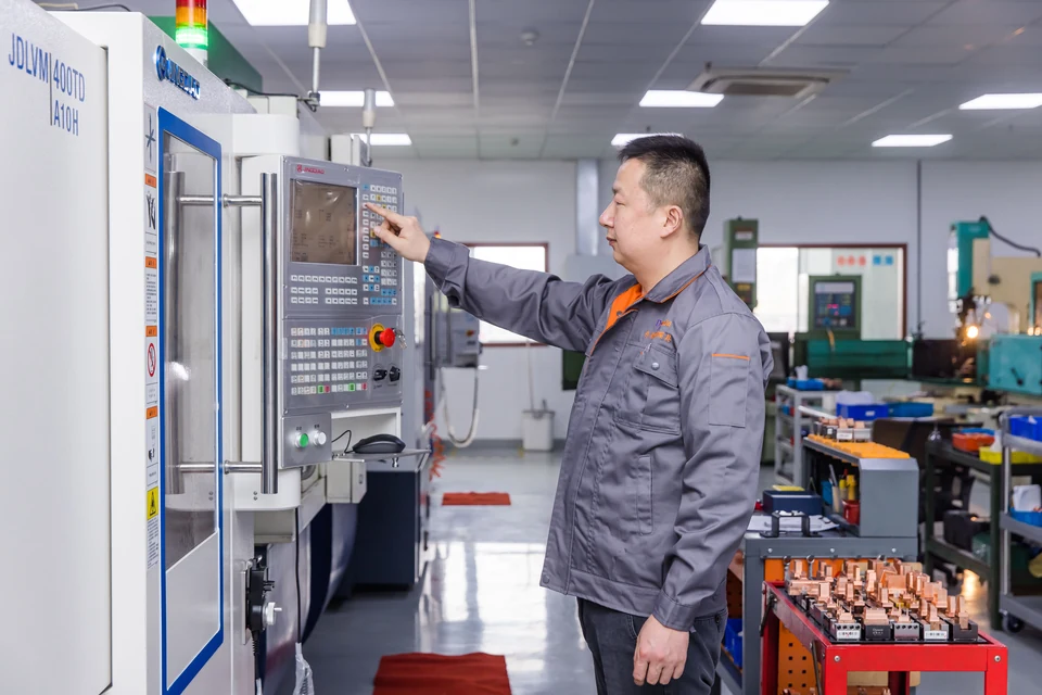 A worker at a CNC machine control panel, with tooling workpieces on a nearby trolley